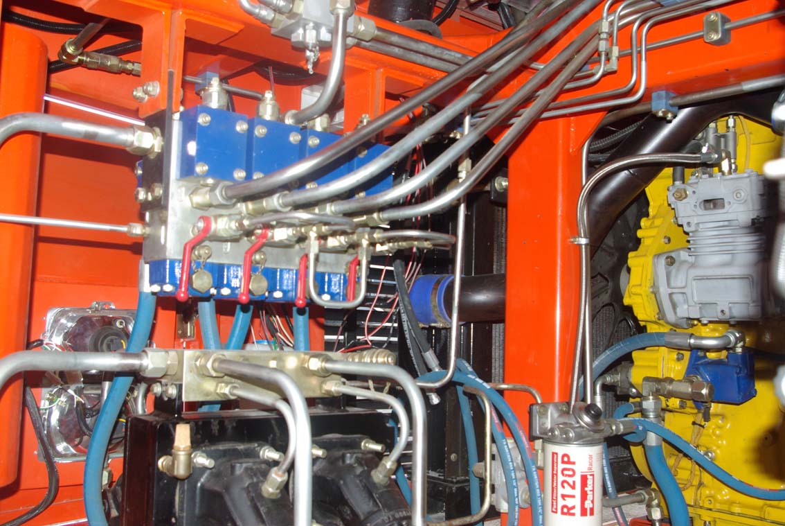 Close-up of a complex hydraulic system in an orange AVAILABLE PALING HEAVY TRANSPORTER, showing metal pipes, valves, blue and red connectors, and a yellow engine component.