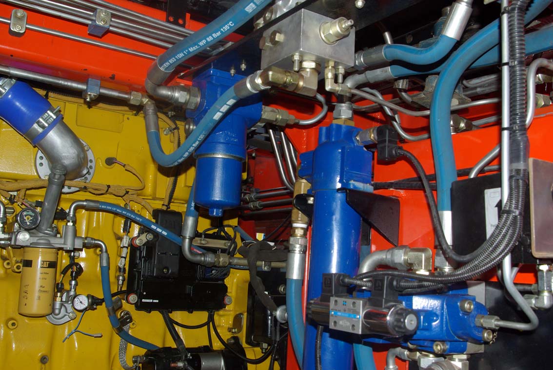 Close-up of the AVAILABLE PALING HEAVY TRANSPORTER at work, highlighting blue hydraulic cylinders, metal pipes, hoses, and yellow and red panels in a complex mechanical setup.