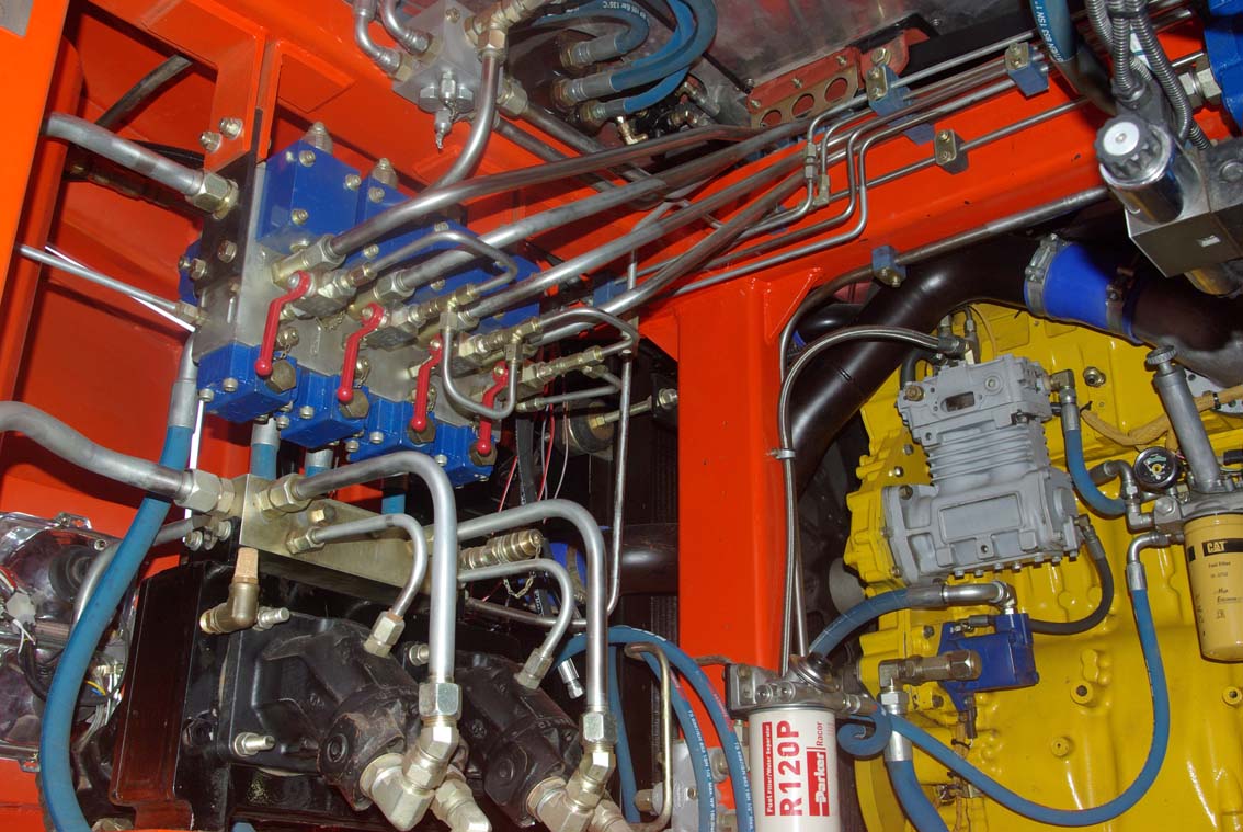 Close-up of a complex hydraulic system in the AVAILABLE PALING HEAVY TRANSPORTER, showing metal pipes, hoses, valves, and a yellow engine part within a red frame. Connectors, filters, and pump parts are also visible.