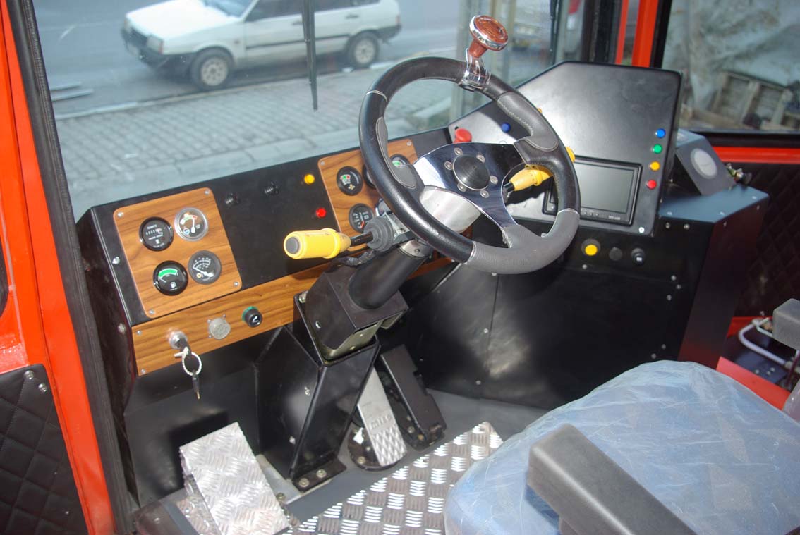 Driver’s seat and control panel of the AVAILABLE PALING HEAVY TRANSPORTER, featuring steering wheel, gauges, switches, key ignition, digital display, metal flooring, padded seat, and a street view through the cab window.