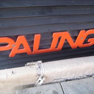 Bold red, 3D letters spell “PALING” on a ribbed black background, evoking the Paling Carrier Iron Mill’s industrial style. Below, a wrapped, damaged section rests on gray—representing the AVAILABLE PALING HEAVY TRANSPORTER.