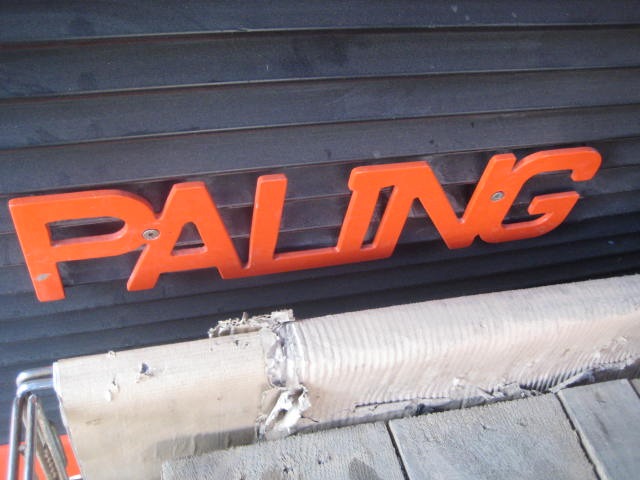 Bold red, 3D letters spell “PALING” on a ribbed black background, evoking the Paling Carrier Iron Mill’s industrial style. Below, a wrapped, damaged section rests on gray—representing the AVAILABLE PALING HEAVY TRANSPORTER.