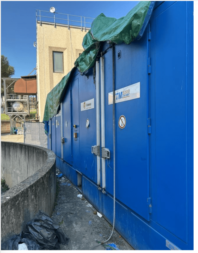 Large blue industrial containers covered partly with a green tarp are positioned outdoors next to a building. Signs with logos and warning symbols are visible on the containers, and some trash is on the ground nearby.