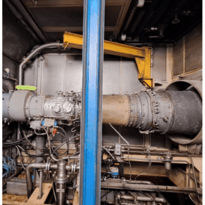 A close-up view of industrial machinery inside a facility, featuring a large metal cylindrical component with pipes, wires, and bolts, partially framed by a blue vertical metal support.