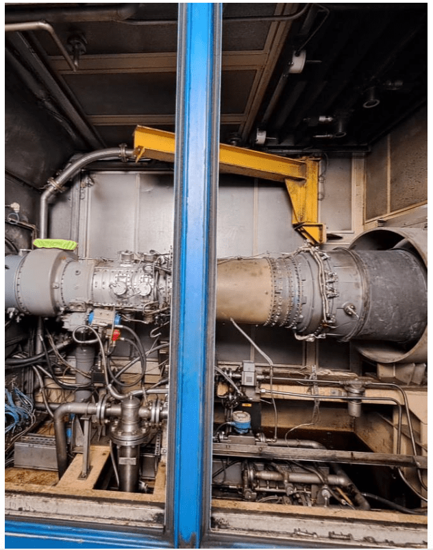 A close-up view of industrial machinery inside a facility, featuring a large metal cylindrical component with pipes, wires, and bolts, partially framed by a blue vertical metal support.