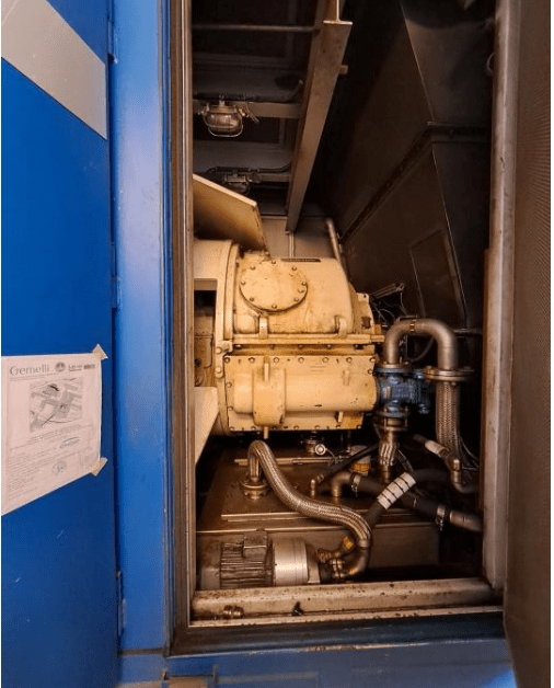 A close-up view of industrial machinery inside a blue metal enclosure, showing pipes, valves, and fittings connected to a large yellow mechanical component. A paper with diagrams is attached to the door.