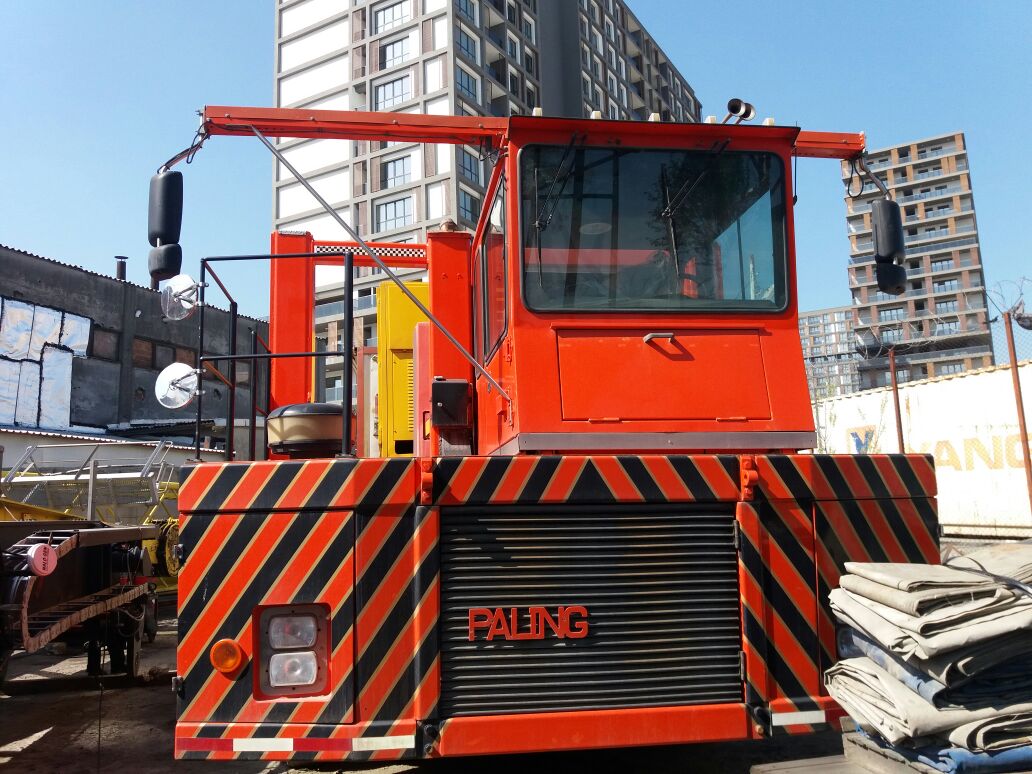 A large red industrial vehicle with black diagonal stripes on the front is parked outdoors near buildings. The sky is clear and there are some construction materials nearby. The word PALFINGER is visible on the vehicle.