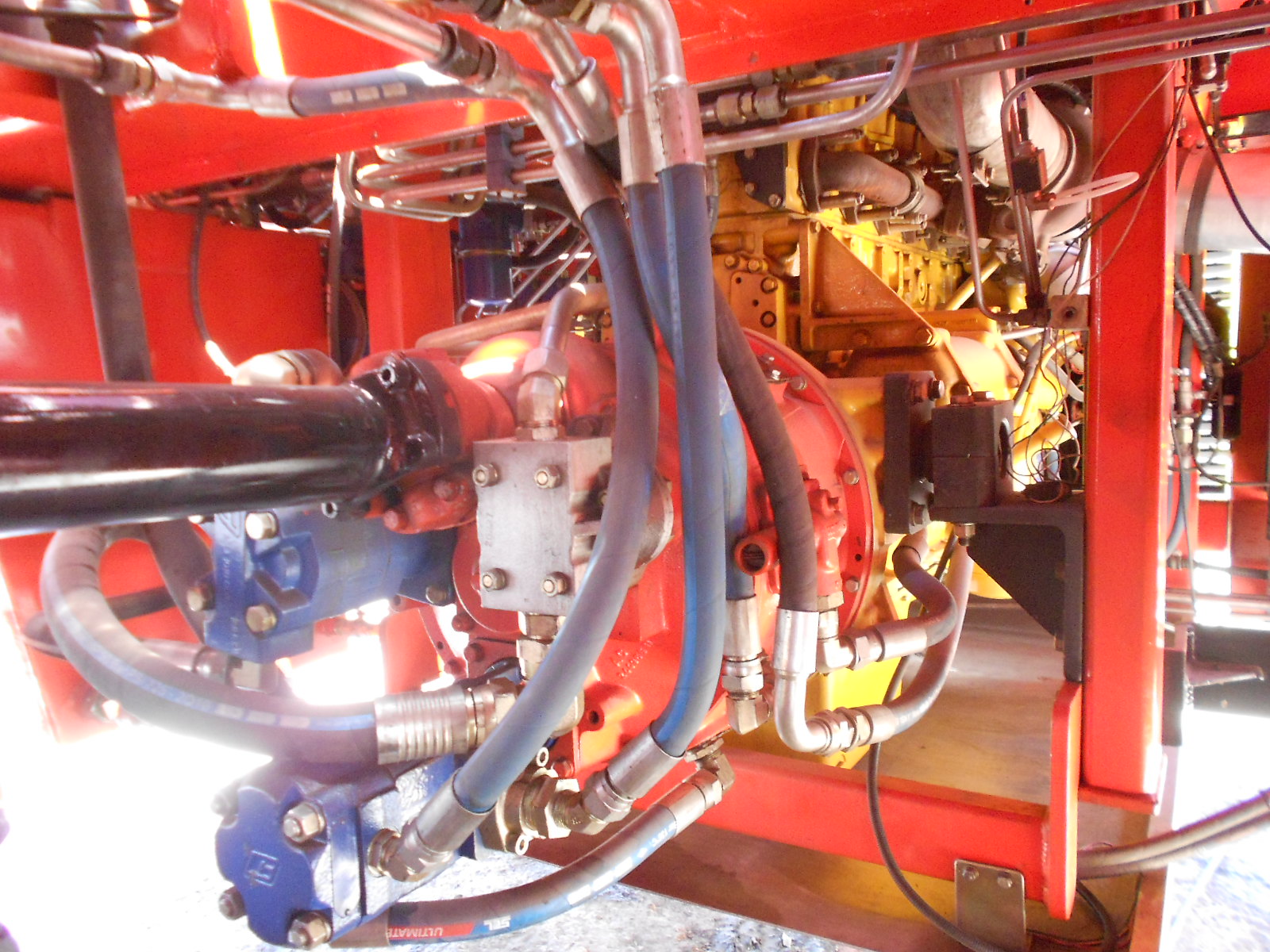 Close-up view of a complex machinery system showing various pipes, hoses, metal components, and a hydraulic pump within a red metal frame. The machinery appears industrial and is brightly lit.
