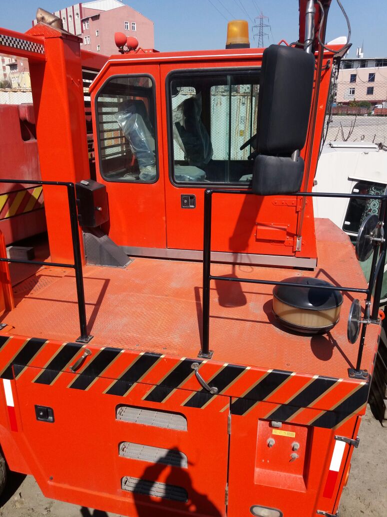 Bright orange industrial vehicle with black and yellow hazard stripes, a black chair mounted near the cabin, safety railings, and a yellow warning light on top. Urban buildings are visible in the background.