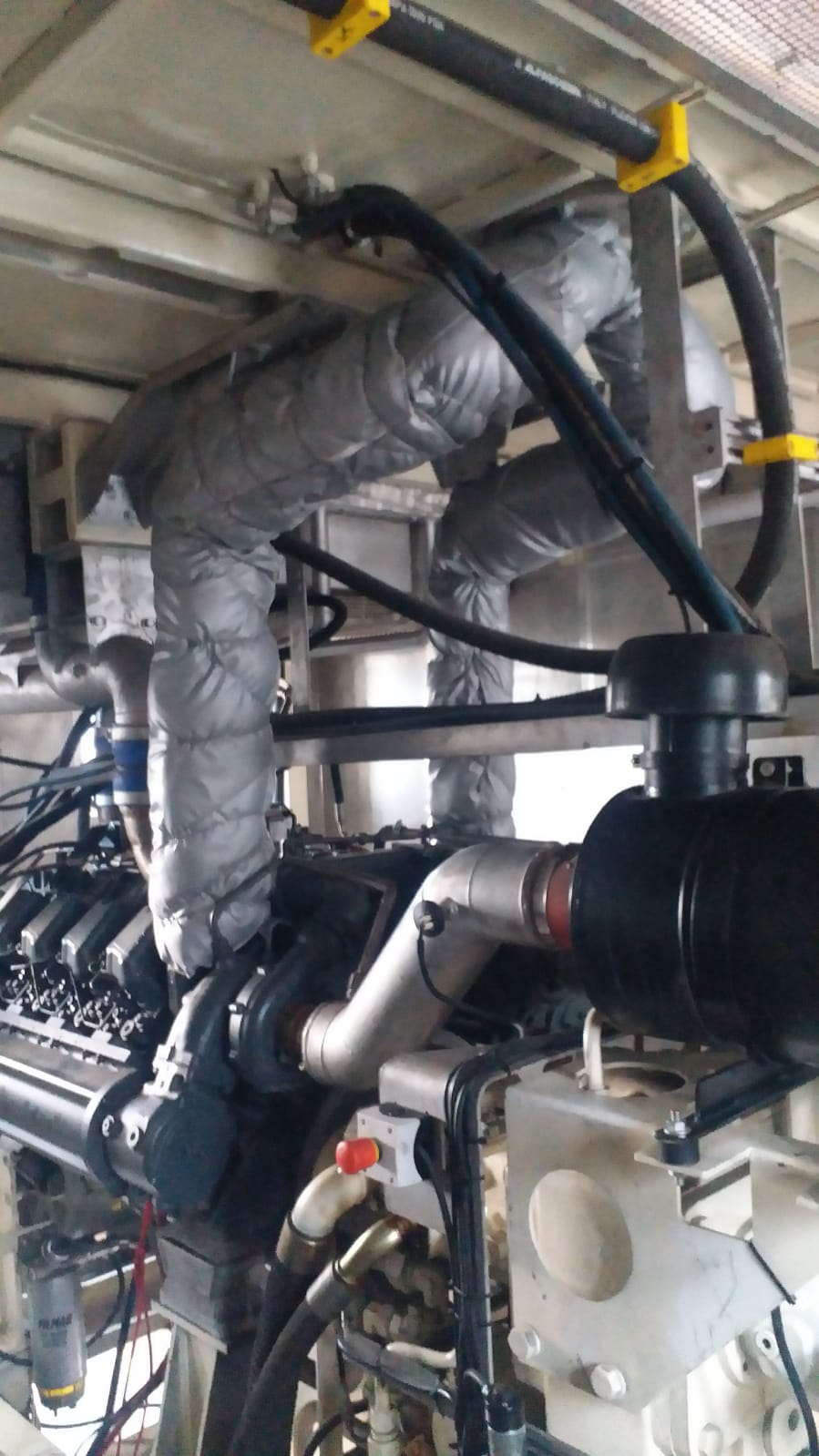 A close-up of the AVAILABLE LIEBHERR MOBILE HARBOUR CRANE LHM150 industrial engine shows pipes, hoses, and insulated exhaust manifolds with gray wrapping inside a metal enclosure.