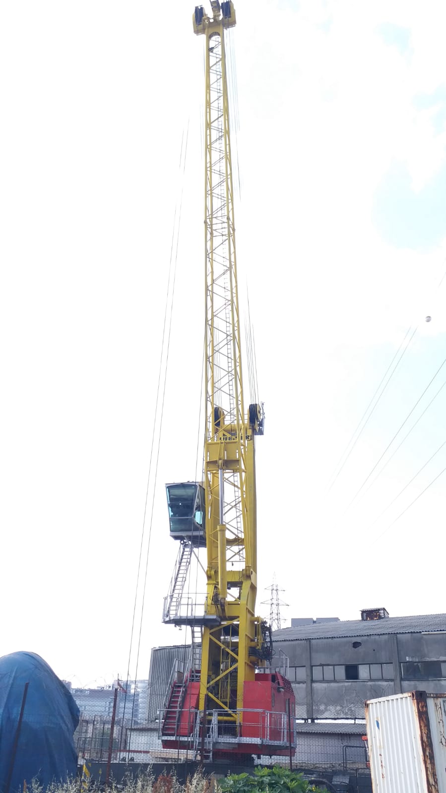 An AVAILABLE LIEBHERR MOBILE HARBOUR CRANE LHM150, tall and yellow with a red base and operator cabin, stands in an industrial area amid buildings, a covered object, and a white container under a mostly clear sky.