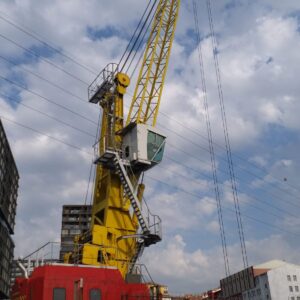 A large yellow crane with a red base stands at a construction site, surrounded by buildings under a partly cloudy sky. Power lines are visible in the background.