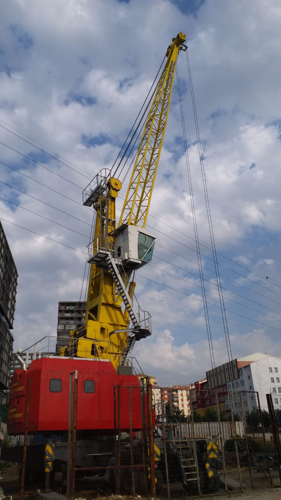 A large yellow crane with a red base stands at a construction site, surrounded by buildings under a partly cloudy sky. Power lines are visible in the background.
