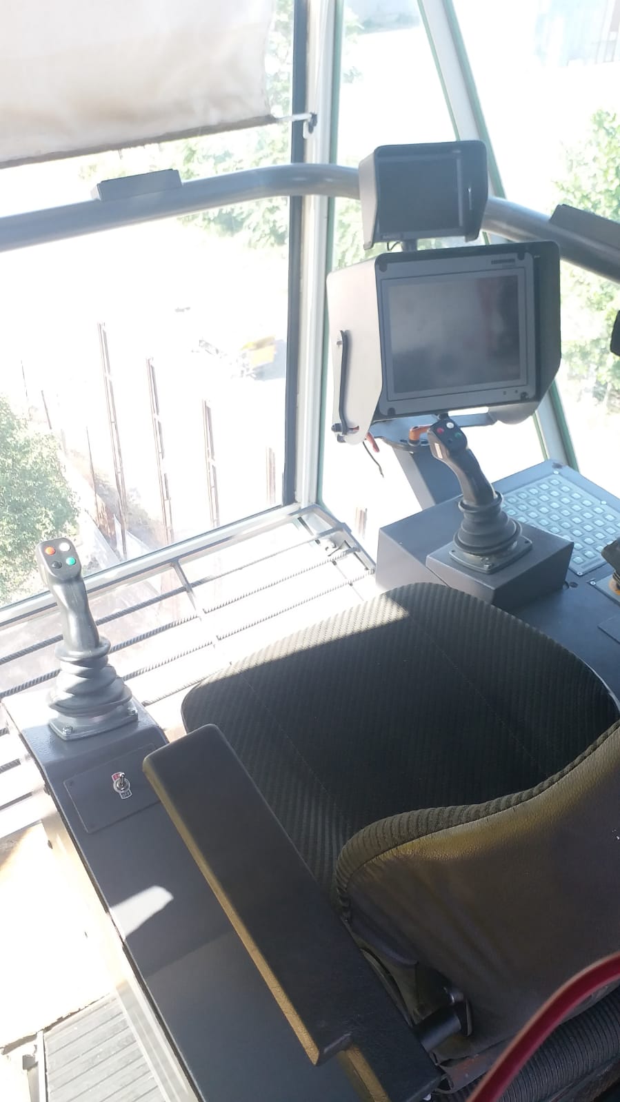 The AVAILABLE LIEBHERR MOBILE HARBOUR CRANE LHM150 features a modern operator’s seat with dual joystick controls and a monitor in a sunlit, glass-enclosed cabin overlooking an outdoor industrial area.