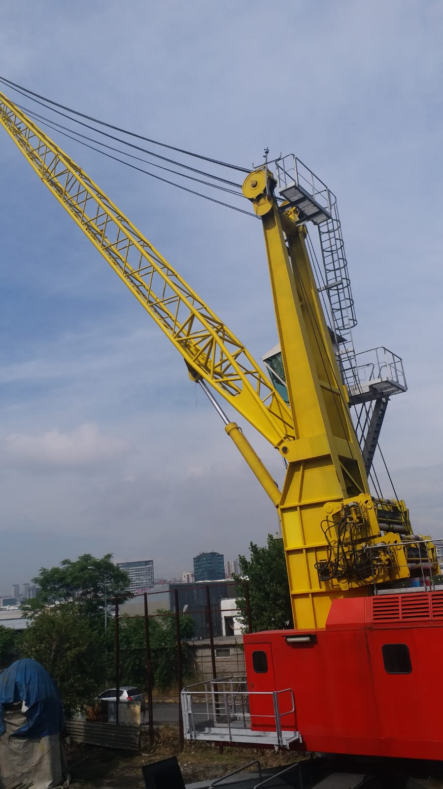 The AVAILABLE LIEBHERR MOBILE HARBOUR CRANE LHM150, featuring a large yellow lattice boom and red base, stands outdoors near trees and buildings under a partly cloudy sky.