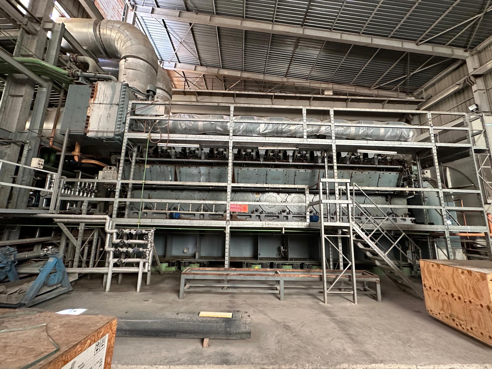 Three used MAN 18V48/60 HFO diesel engines (50 Hz) power plant sets are displayed in a spacious, well-lit factory with metal scaffolding, pipes, and various equipment beneath a corrugated metal roof.