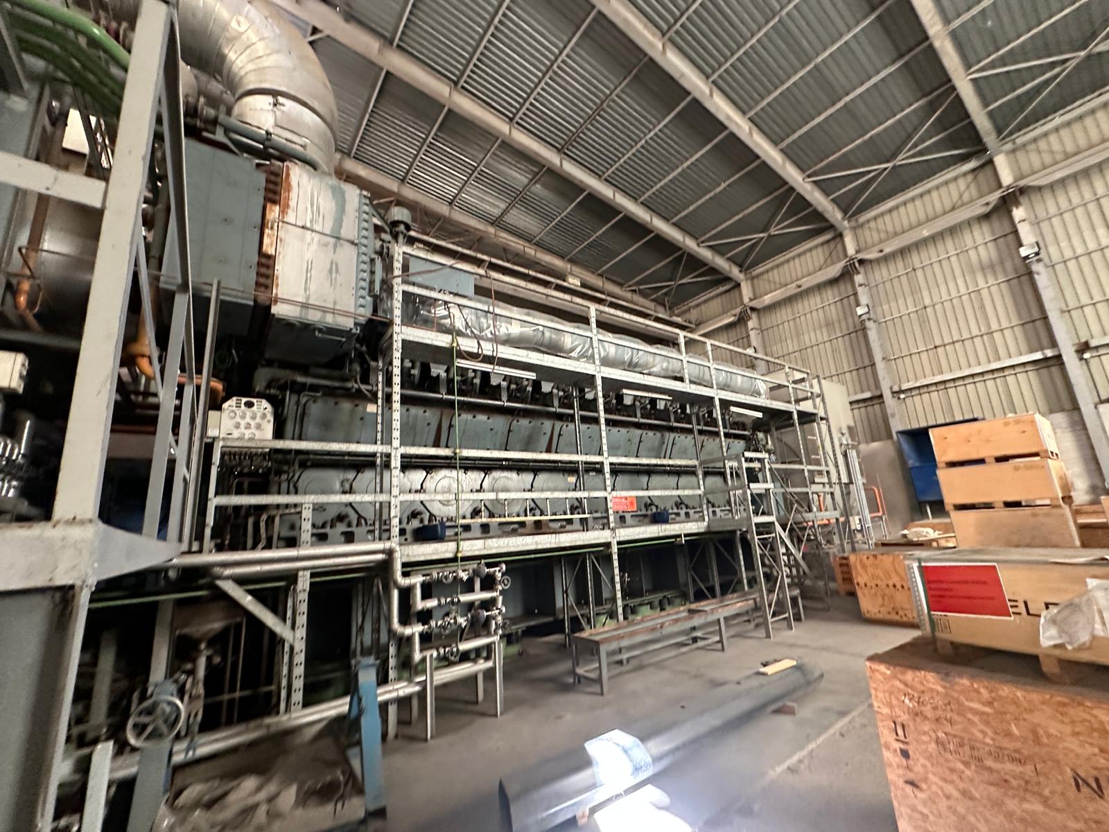A spacious warehouse with metal walls and a high ceiling contains 3 sets of used 18V48/60 MAN HFO diesel engines (50 Hz power plant), surrounded by pipes, platforms, control panels, and scattered wooden crates.