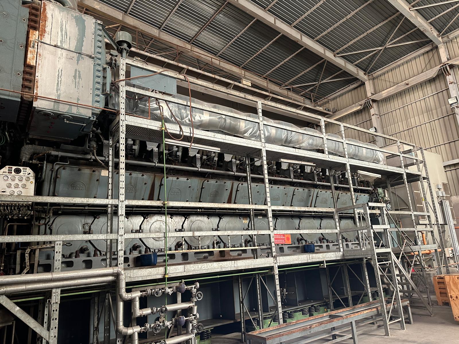 Three sets of used 18V48/60 MAN HFO diesel engines (50 Hz power plant) are installed in a factory, surrounded by metal scaffolding and piping beneath a high metal roof with beams and corrugated panels.