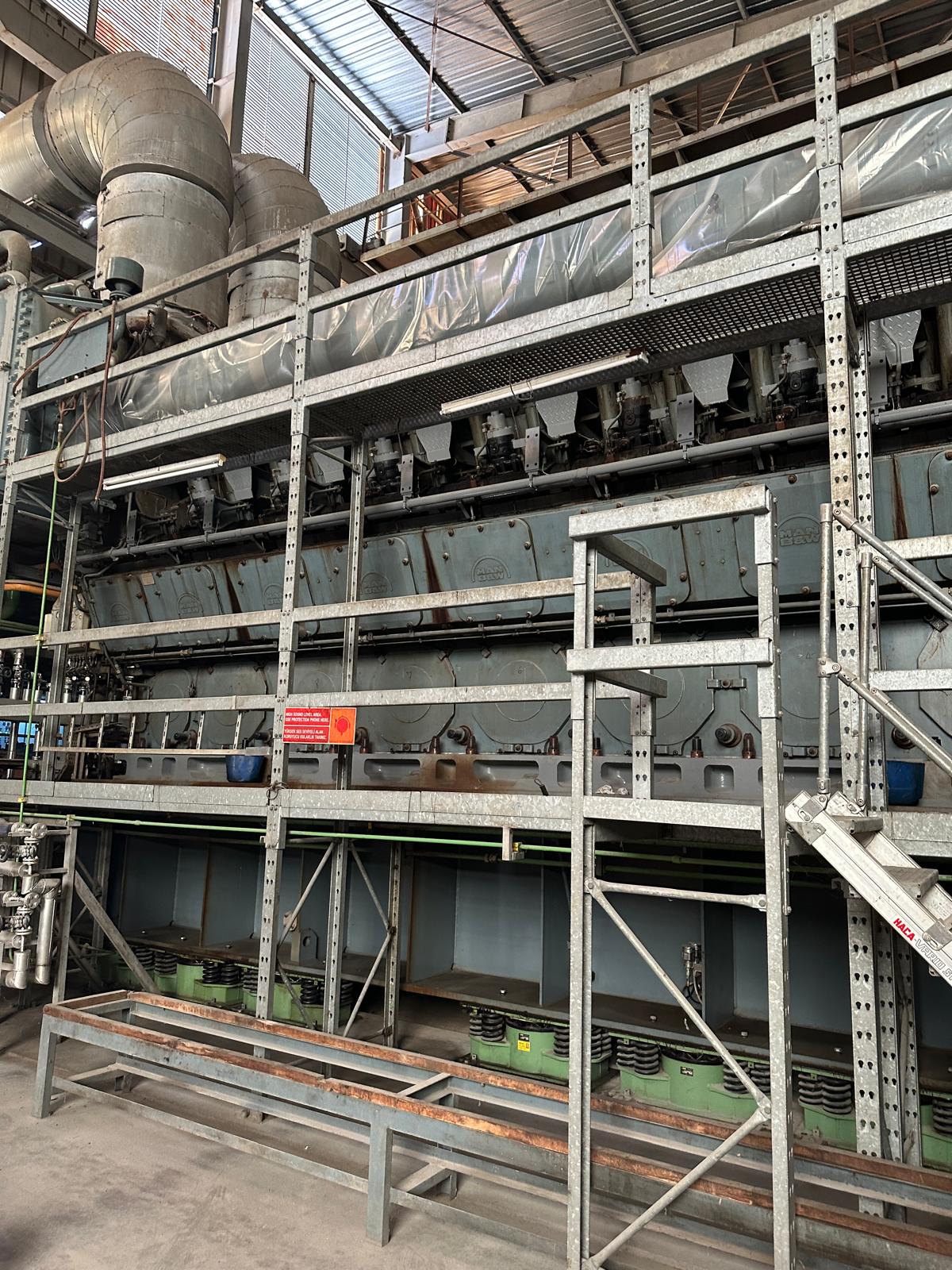 A spacious HFO power plant houses 3 sets of used 18V48/60 MAN diesel engines (50 Hz), featuring metal pipes, control panels, and a robust metal framework beneath a high factory ceiling.
