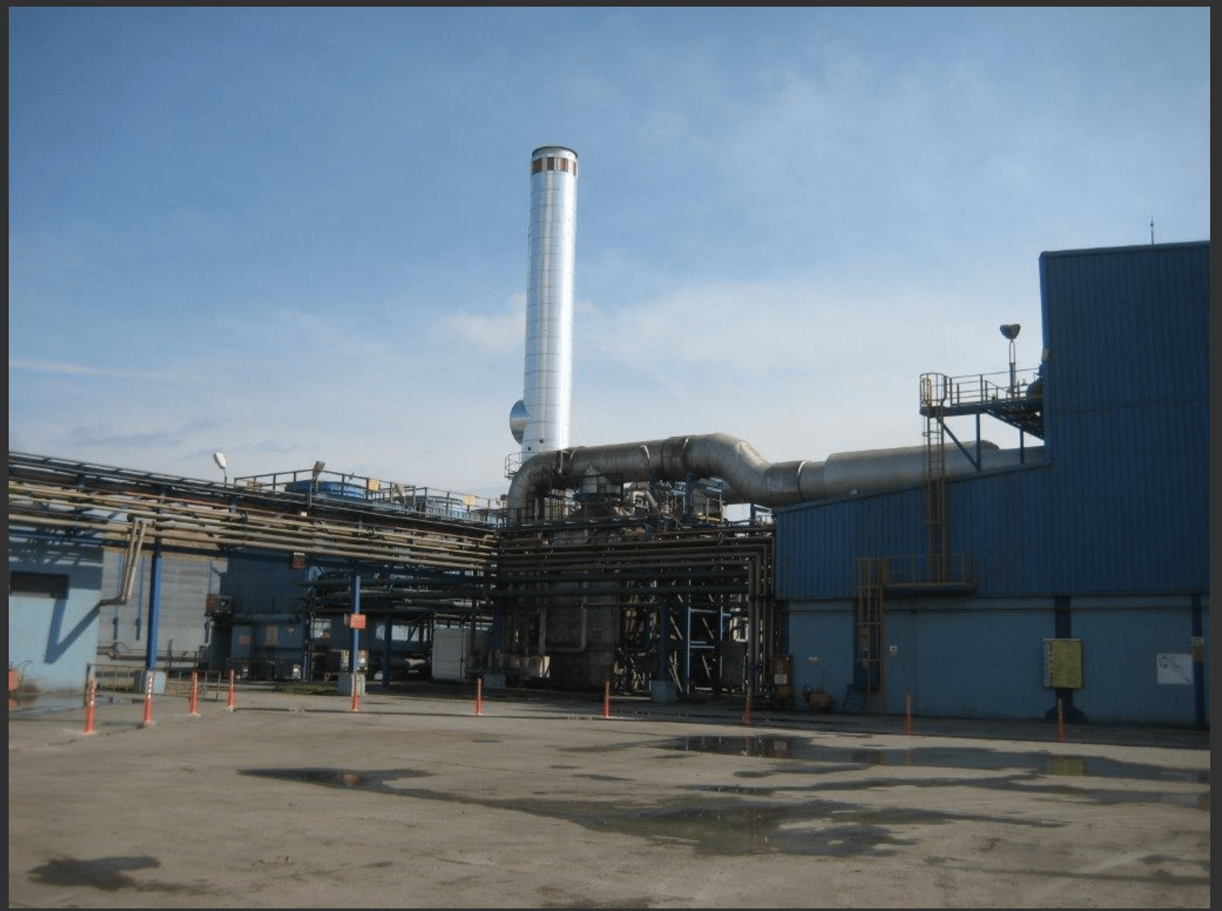 A power plant featuring 3 sets of used 18V48/60 MAN HFO diesel engines (50 Hz), with blue buildings, a tall white smokestack, metal pipes and structures. Concrete ground shows puddles and orange traffic cones; sky is mostly clear.
