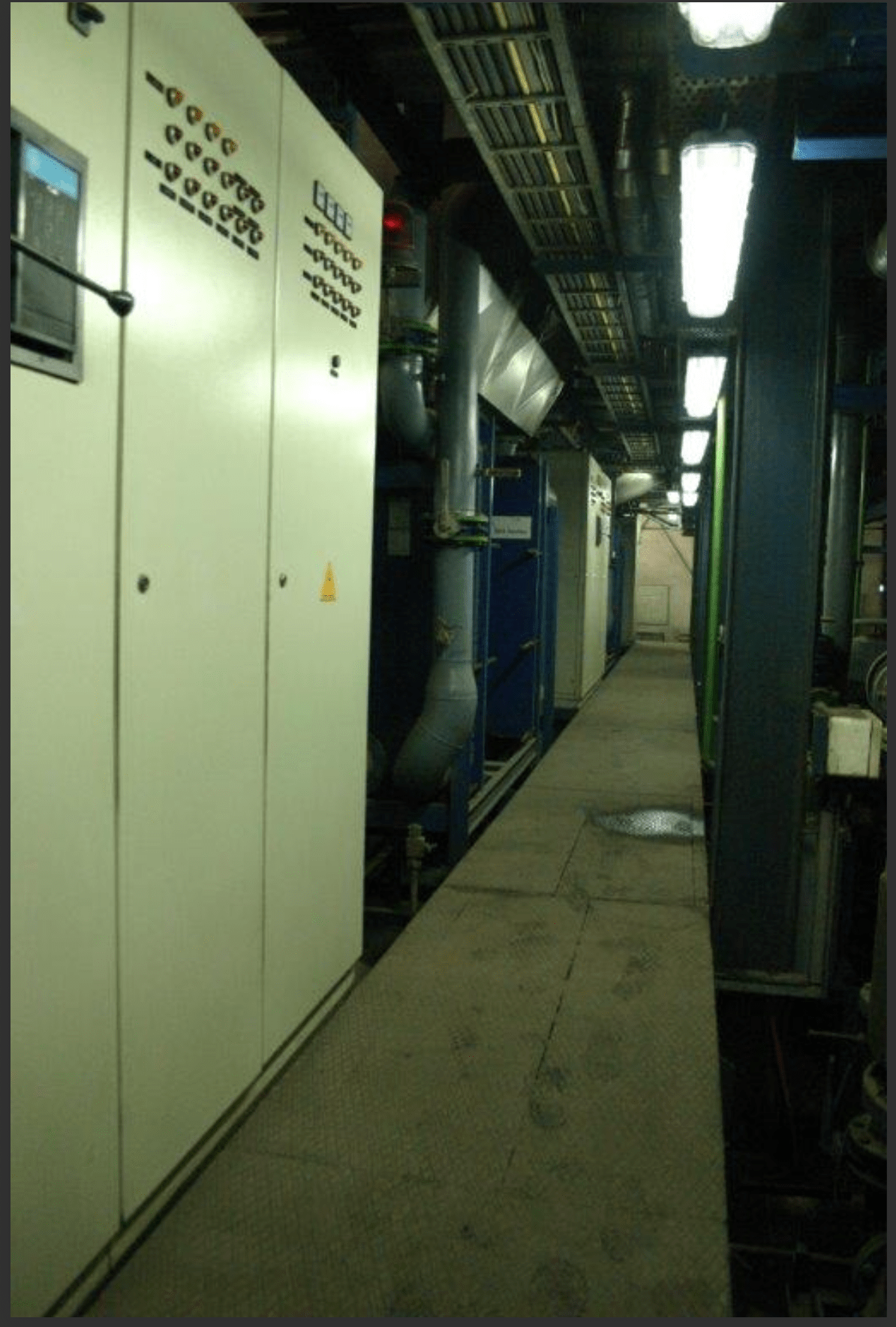 A narrow industrial corridor in a 3 SETS OF USED 18V48/60 MAN HFO DIESEL ENGINES 50 HZ POWER PLANT, with control panels left, blue machinery right, overhead lighting, and a grated metal walkway down the center.