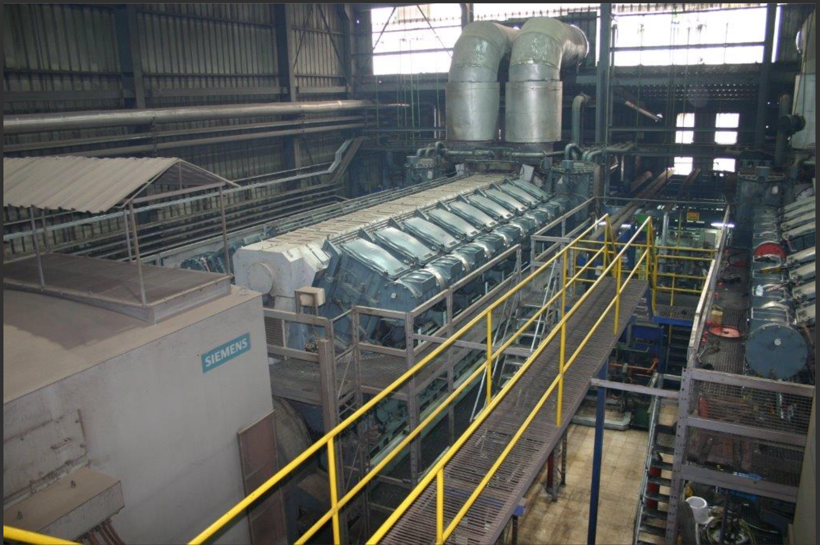 An industrial facility interior features large machinery, including 3 sets of used 18V48/60 MAN HFO diesel engines (50 Hz power plant), metal walkways with yellow railings, pipes, ducts, and various equipment in a spacious, well-lit space.