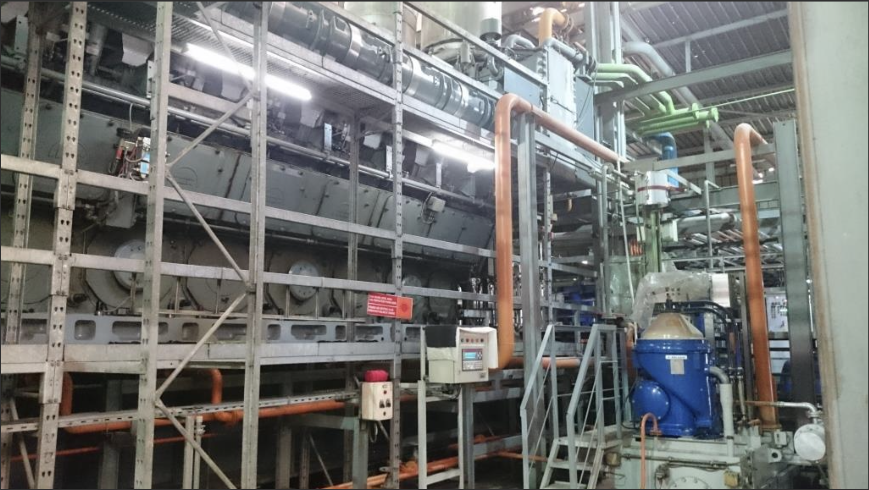 Inside a factory, large industrial machinery features metal pipes, railings, and control panels. Notably, three used 18V48/60 MAN HFO diesel engines (50 Hz) display blue cylindrical units with orange pipes and cables under fluorescent lighting.
