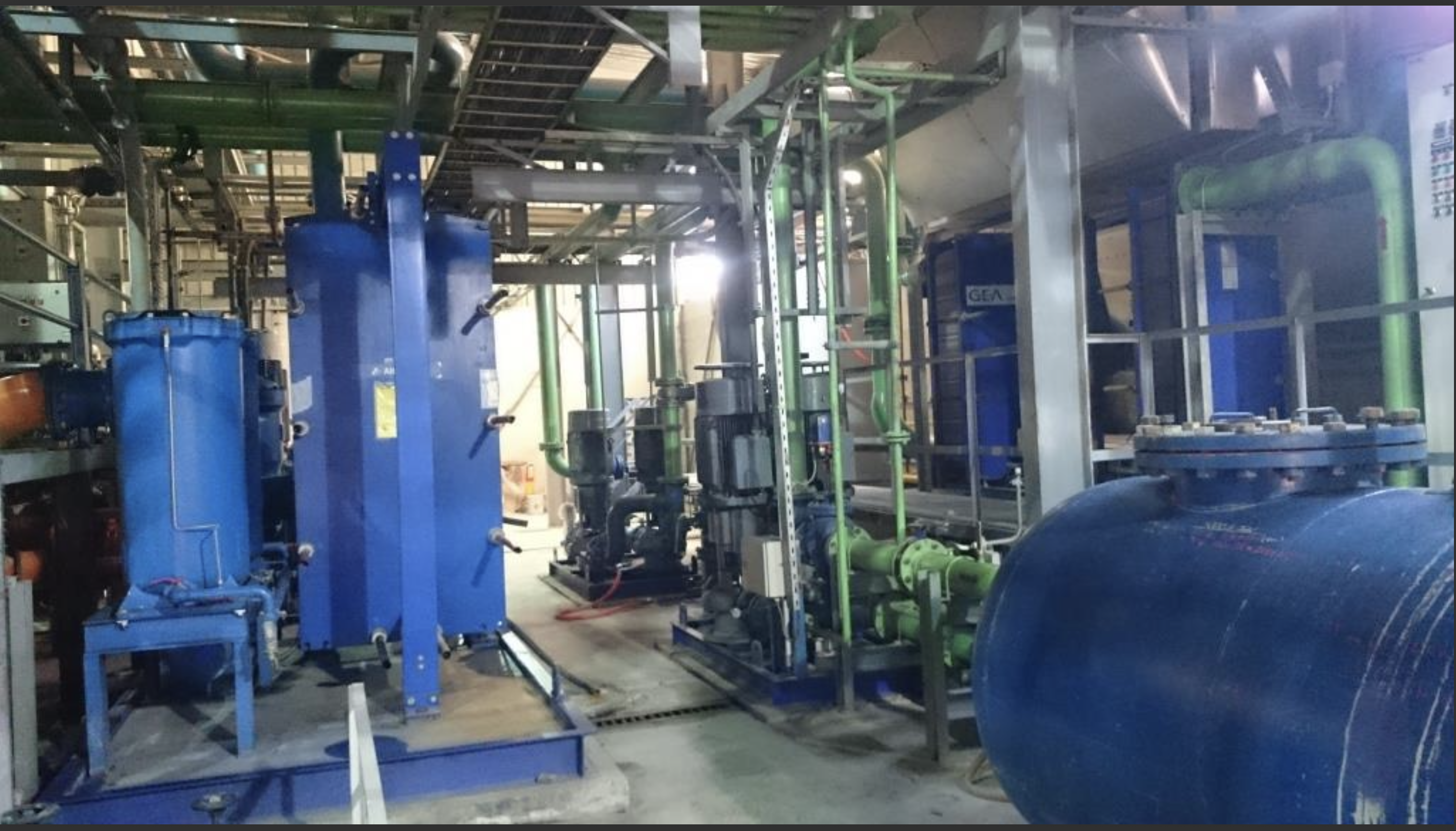 Interior of a power plant featuring 3 sets of used 18V48/60 MAN HFO diesel engines (50 Hz), with large blue tanks, green and metal pipes, valves, and structural components illuminated by bright overhead lighting.