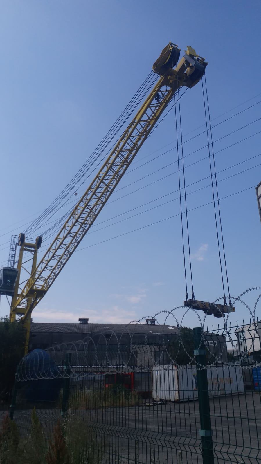 A large yellow crane lifts materials high above a fenced industrial area with barbed wire, shipping containers, and warehouses on a clear day with blue sky.
