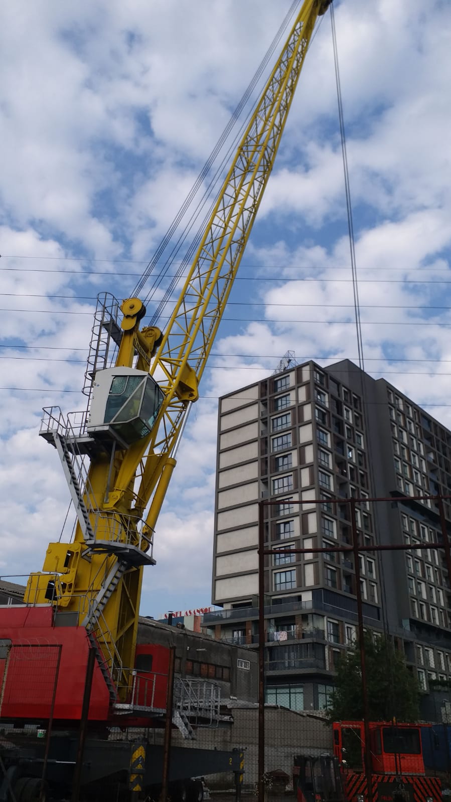 A yellow crane with a glass cabin stands in front of a modern multi-story building under a partly cloudy sky, with power lines visible overhead.