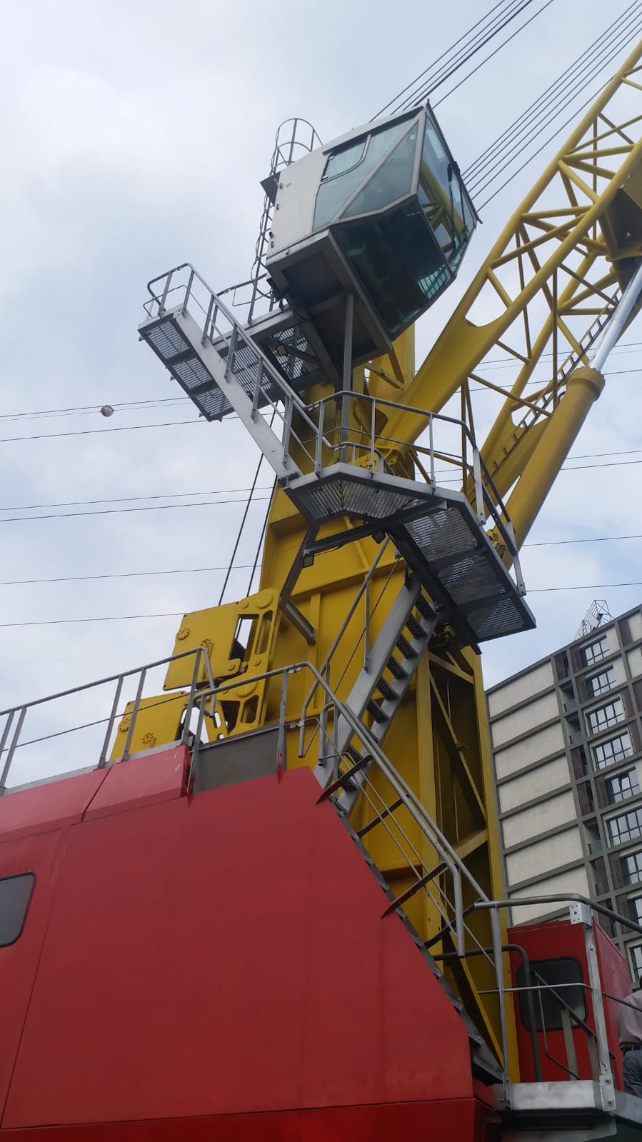 A close-up of the AVAILABLE LIEBHERR MOBILE HARBOUR CRANE LHM150 shows its large yellow frame, glass control cabin, metal stairs and railings, with a red lower section. A building and cloudy sky appear in the background.