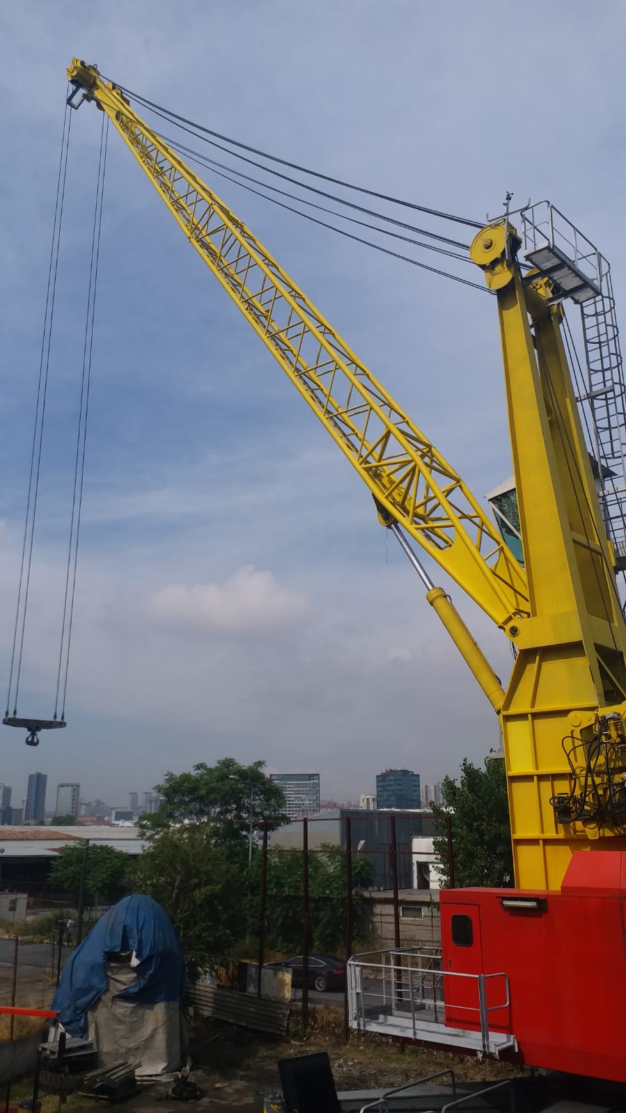 An AVAILABLE LIEBHERR MOBILE HARBOUR CRANE LHM150 with a lattice boom stands outdoors near city buildings, a blue tarp-covered object nearby, under a partly cloudy sky.