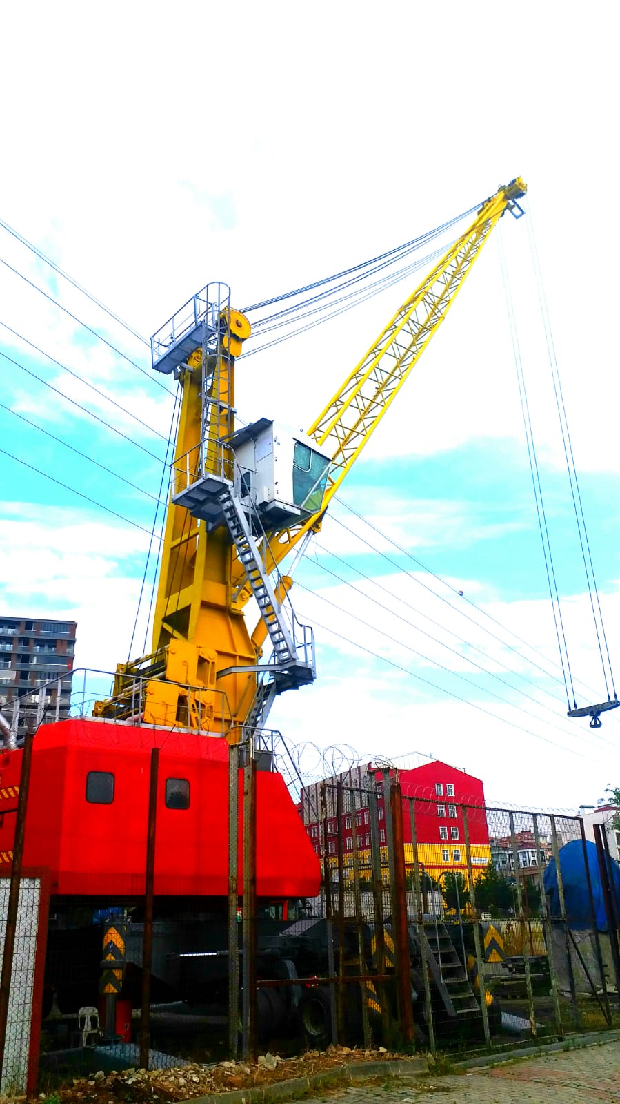 The AVAILABLE LIEBHERR MOBILE HARBOUR CRANE LHM150, in yellow and red, stands behind a metal fence with buildings and a cloudy sky behind it. Power lines cross the view.