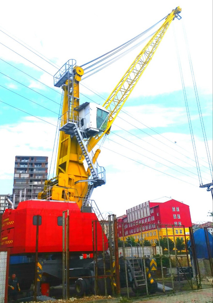 A large, colorful dockside crane with a red base and a long yellow boom stands near a fence, with apartment buildings and a red warehouse in the background under a partly cloudy sky.