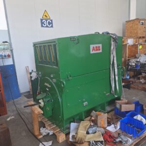 A large green industrial machine labeled ABB is placed on a wooden pallet in a workshop, surrounded by tools, boxes, gloves, cables, and other equipment. A 3C sign is on the wall behind it.