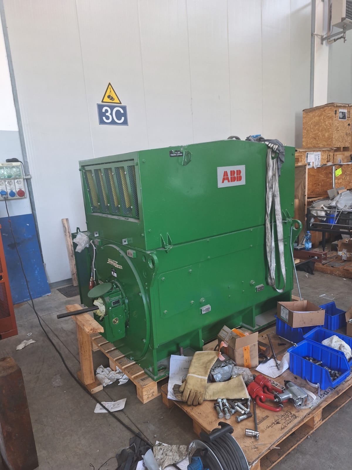 A large green industrial machine labeled ABB is placed on a wooden pallet in a workshop, surrounded by tools, boxes, gloves, cables, and other equipment. A 3C sign is on the wall behind it.