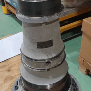 A large metallic turbine engine component with multiple rings and bolts is standing upright on a wooden table in an industrial setting. Shelving and boxes are visible in the background.