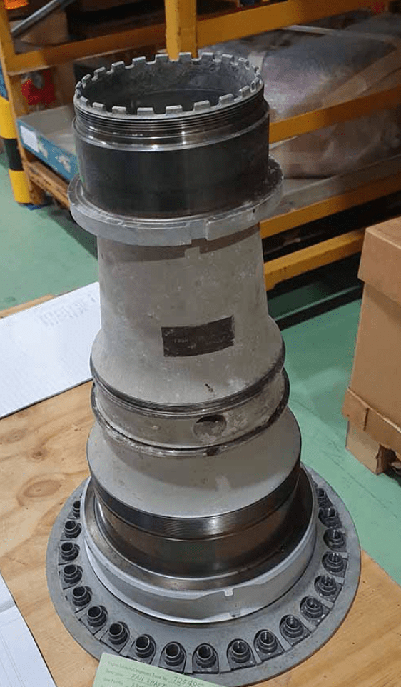 A large metallic turbine engine component with multiple rings and bolts is standing upright on a wooden table in an industrial setting. Shelving and boxes are visible in the background.