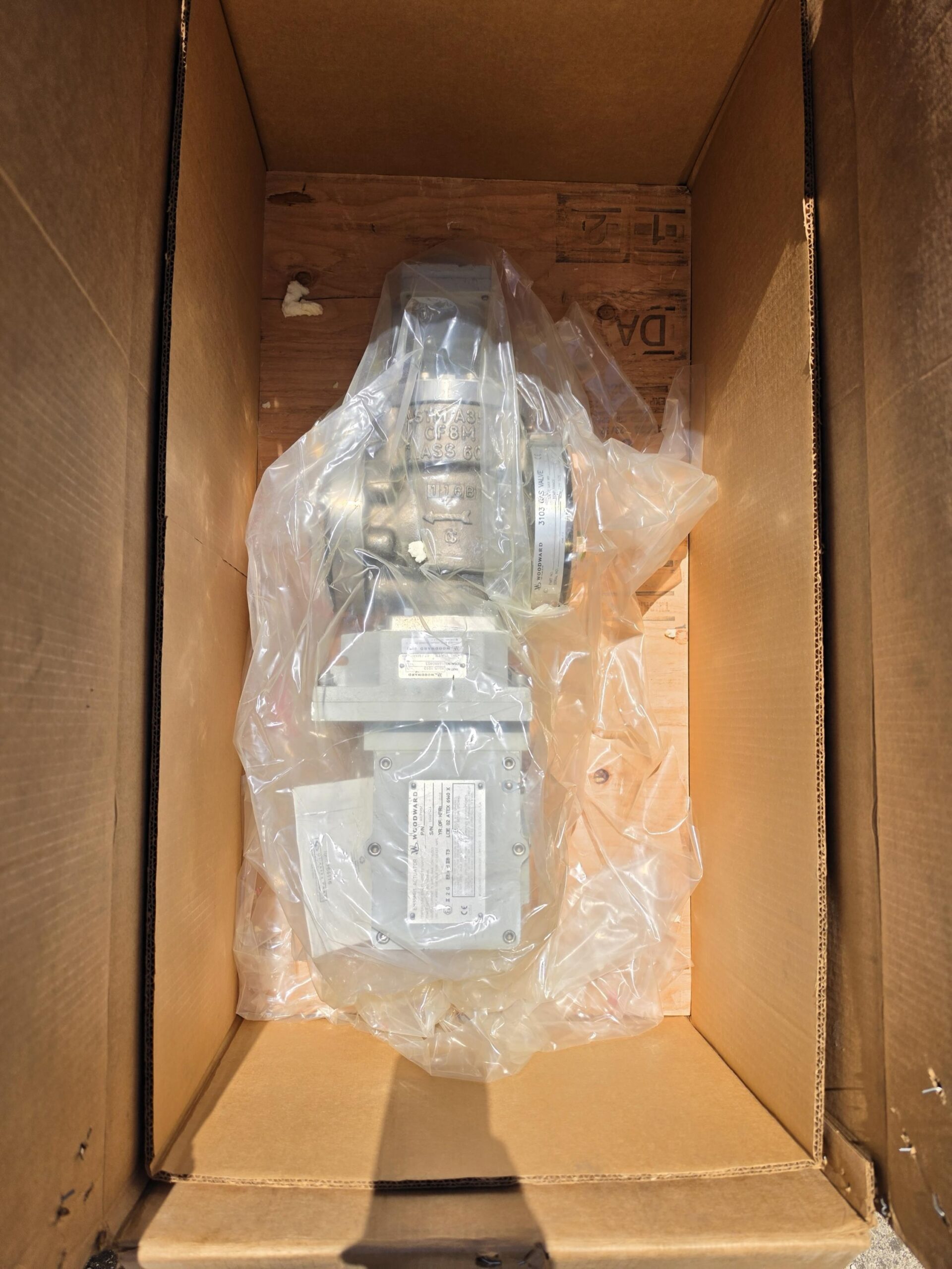 A metal industrial component wrapped in clear plastic is placed inside a large cardboard box lined with wooden panels. Sunlight casts shadows on the box walls.