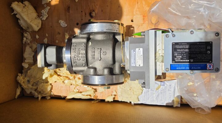Woodward Gas Valves (PN 8915-1310), new and never used or installed, sit in a cardboard box with labels attached. The valves are surrounded by yellow packing foam and plastic for protection, typical of components shipped for heavy industry or gas turbine maintenance. Visible paperwork beneath them suggests these parts are intended for an LM2500 engine assembly. Sunlight casts shadows across the box and its contents.