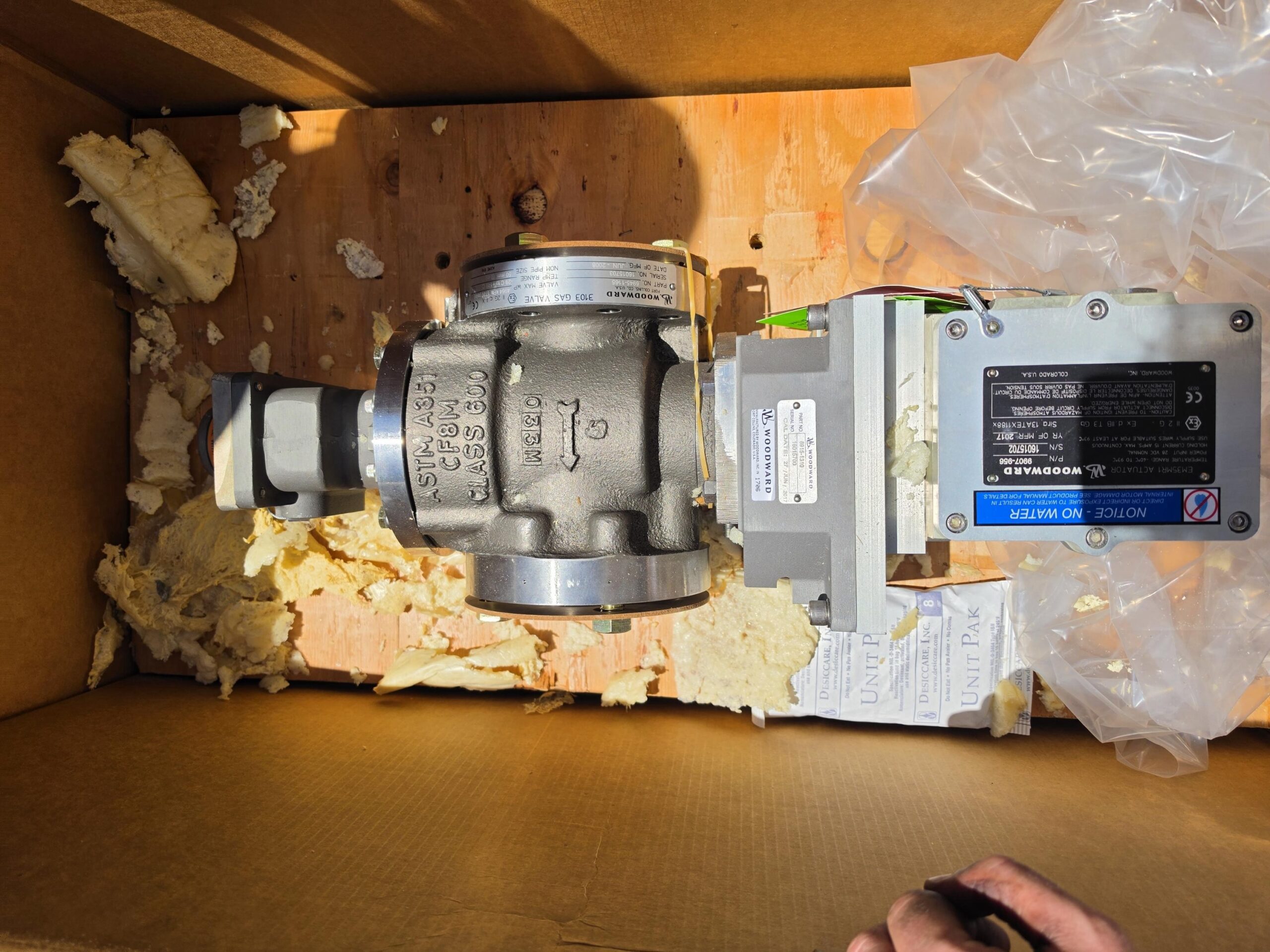 A metal valve with attached electronic components sits in a cardboard box lined with foam pieces and plastic, with some paperwork visible underneath. Sunlight casts a shadow on the box.