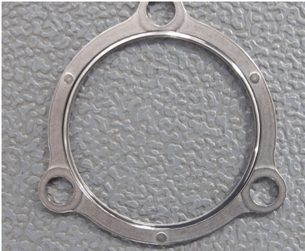 The AVAILABLE 9559M20G01 RING ASSY, SEAL NOZZLE GASKET—a metallic circular gasket with three extended holes—is placed on a textured gray surface.
